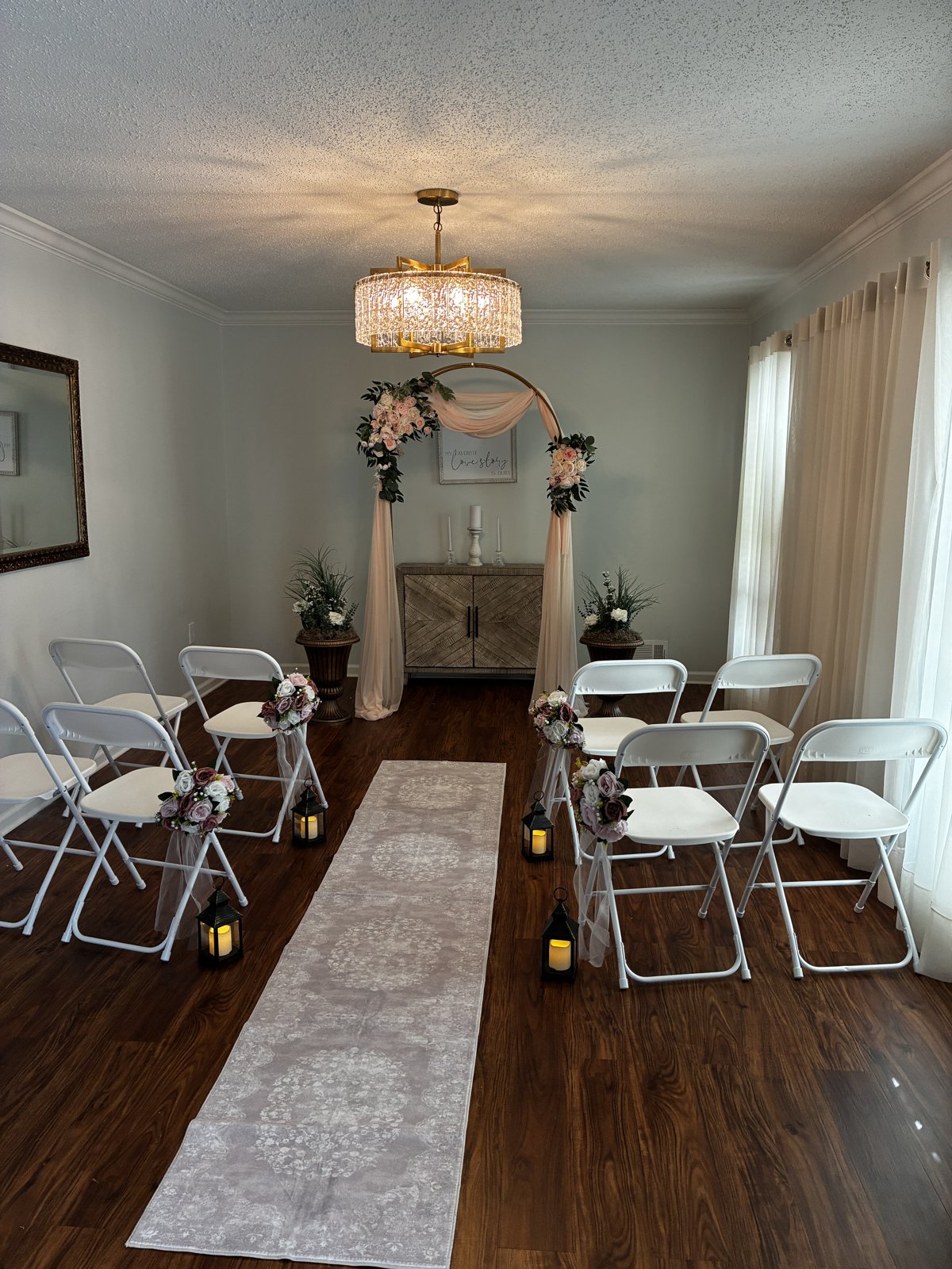 The ceremony room set for a wedding, with a blush draped gold arch under a crystal chandelier, white chairs on both sides of a runner, and lanterns along the aisle